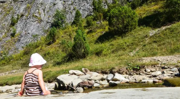 Dziecko w kapelusiku siedzi nad strumieniem w Małych Pieninach, otoczone zielenią i skalistym zboczem.