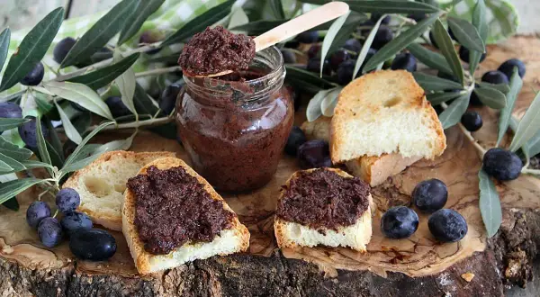 Ecco come si fa il patè di olive: bruschette con paté scuro, olive nere e rametti d'ulivo su un tagliere di legno.
