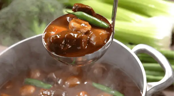 A ladle scoops tender, slow-cooked meat in rich gravy from a steaming pot. Perfect for learning jak dusić mięso.
