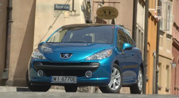 Błękitny Peugeot 207 z wyraźnymi żarówkami reflektorów stoi na brukowanej ulicy.