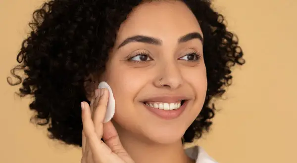 Donna sorridente con capelli ricci usa un dischetto per struccarsi, mostrando come si usa il burro struccante per una pelle luminosa.