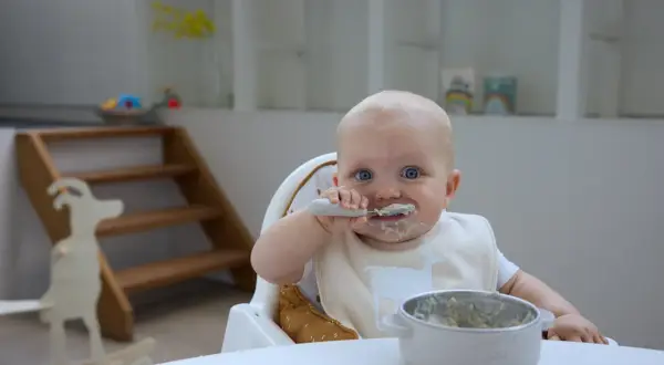 Ein süßer **Joghurt Baby** mit blauen Augen isst mit Löffel. Der Latz ist mit einer Ziege verziert.
