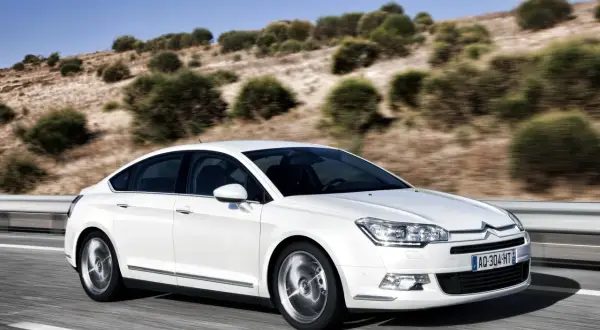 Biały sedan Citroën C5 w ruchu na drodze z pagórkowatym, zadrzewionym krajobrazem w tle.