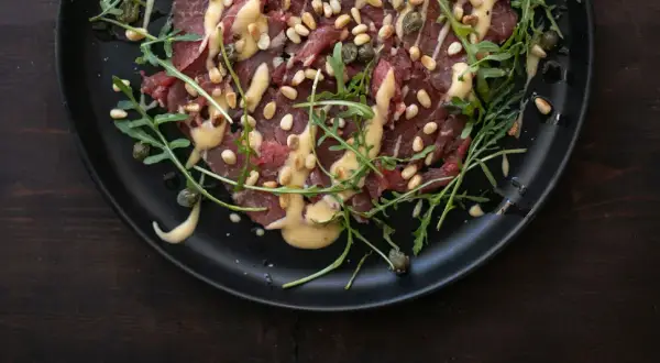 Rinder Carpaccio mit Pinienkernen, Kapern und Rucola auf schwarzer Platte, beträufelt mit einer cremigen Sauce.