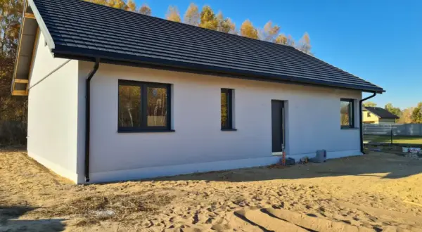 Nowy dom z szarym dachem i białymi ścianami, z oknami i drzwiami, w stanie surowym zamkniętym.