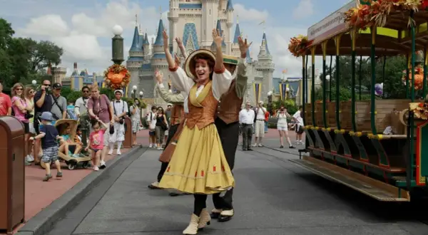 Uczestnicy parady w Disneylandzie pozdrawiają tłum. W tle zamek, a obok zabytkowy tramwaj.