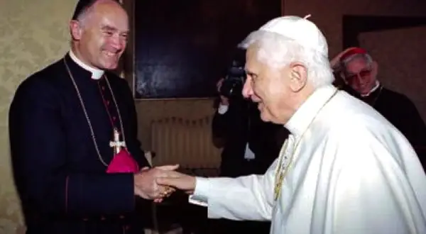 Papst Benedikt XVI. schüttelt einem Bischof die Hand. Im Hintergrund ein Kameramann und ein weiterer Geistlicher, ein Zeichen der Würde der summorum pontificum.