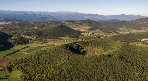 Paisaje de la ruta volcanes Garrotxa, con conos volcánicos cubiertos de bosque y campos verdes, bajo un cielo azul.