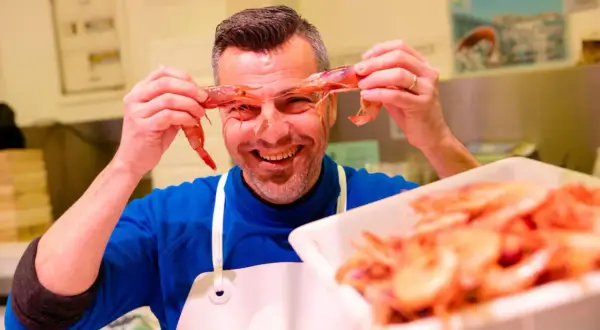 Hombre sonriente jugando con mariscos rojos, sosteniendo dos gambas frente a sus ojos.