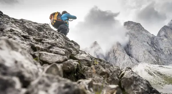 Un randonneur avec un grand sac à dos escalade un sentier rocheux, les pics des Dolomites se dressent dans le brouillard.