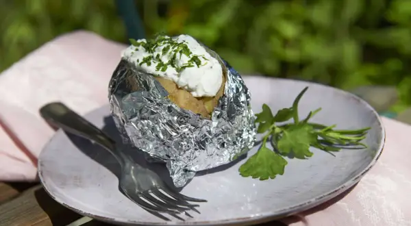 Folienkartoffeln vorkochen: Eine Kartoffel in Alufolie, belegt mit Quark und Kräutern, auf einem Teller mit Gabel und Petersilie.