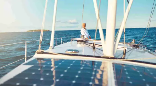 Un uomo sorride sul ponte di una barca a vela, con un impianto pannello solare che riflette il cielo azzurro.