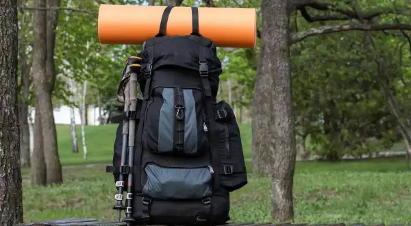 Ein schwarzer Rucksack mit Wanderstöcken und einer orangefarbenen Isomatte, die mit Schlaufen befestigt ist, steht auf einer Holzbank im Grünen.