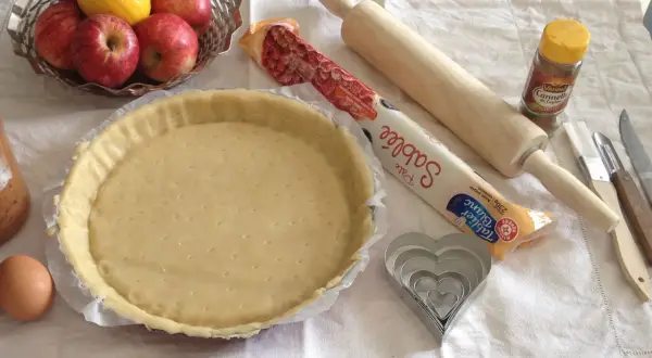 Préparation d'une tarte aux pommes simple : pâte sablée, pommes, citron, œuf, rouleau, emporte-pièces cœur et cannelle.