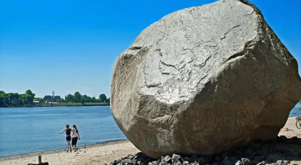 Der alte Schwede, ein riesiger Findling, thront am Ufer. Zwei Frauen spazieren am Wasser entlang, im Hintergrund Bäume und ein blauer Himmel.