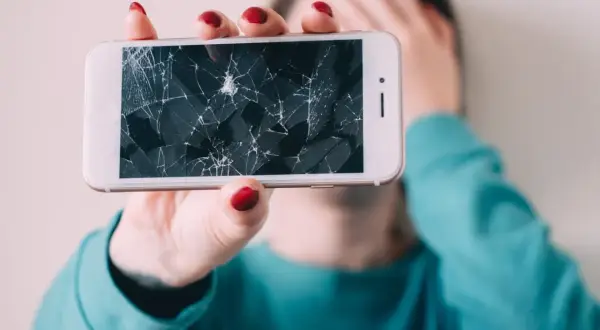 Złamany telefon z pękniętym ekranem, który osoba trzyma przed twarzą, zakrywając ją ręką.