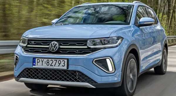 Niebieski VW T-Cross pędzi leśną drogą, odbijając drzewa w szybie.