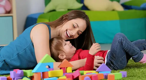 Uśmiechnięta opiekunka bawi się klockami z rocznym dzieckiem. Zastanawiasz się, ile kosztuje niania do rocznego dziecka?