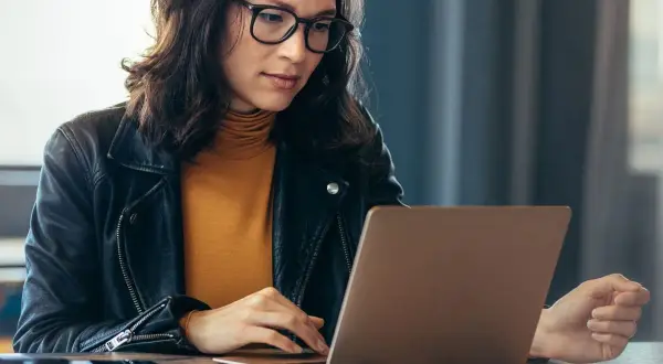 Kobieta w okularach i skórzanej kurtce pracuje na laptopie, szukając informacji, jak sprawdzić lata pracy w ZUS PUE.