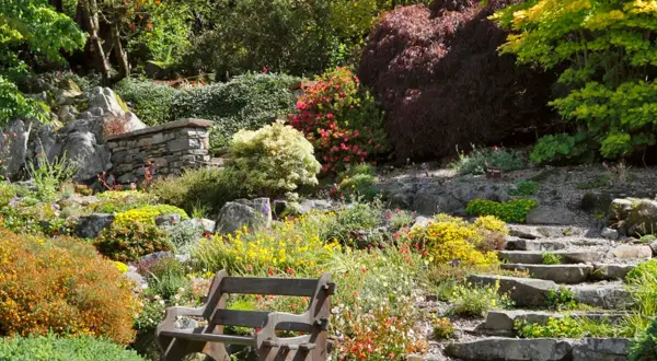 Ein terrassierter Garten am Hang mit Steinwegen und einer Holzbank. Bunte Blumen und Sträucher bedecken die Hänge.