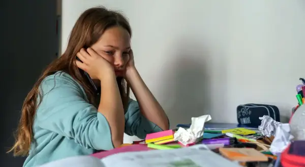 Mädchen sitzt gelangweilt am Schreibtisch, umgeben von Schulmaterialien. Endlich wieder Schule, aber die Motivation fehlt.
