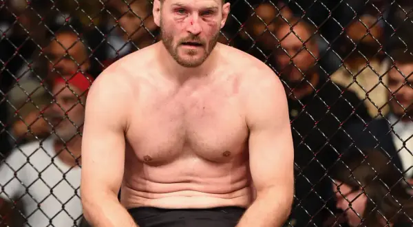 Stipe Miocic, med blodiga sår i ansiktet, sitter utmattad i buren efter en tuff match.