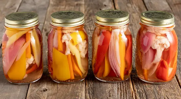 Quattro barattoli di giardiniera fatta in casa, con verdure colorate come peperoni, carote e cavolfiore, su un tavolo di legno rustico.