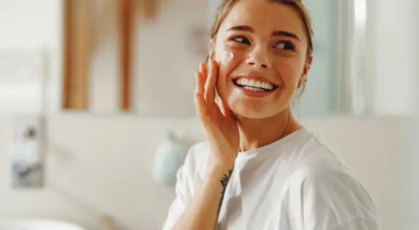 Une femme souriante applique une crème sur sa joue. Elle se prépare pour la journée, peut-être un primer avant ou après crème solaire.