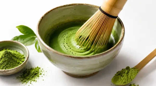 Tradycyjny matcha przepis: bambusowa miotełka ubija zielony proszek w misce, tworząc piankę. Obok miseczka z proszkiem i drewniana łyżeczka.