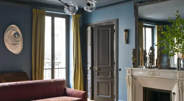Soggiorno elegante con pareti blu balena Pantone, divano bordeaux, tendaggi ocra e camino in marmo.