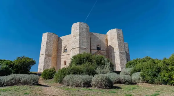 Castel del Monte, un'imponente fortezza ottagonale in pietra chiara, si erge maestoso sotto un cielo azzurro intenso, circondato da vegetazione mediterranea.