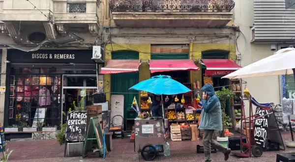Un hombre con capucha camina por una calle adoquinada en Montevideo, pasando por tiendas de discos y un puesto de frutas.