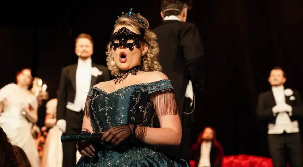 Kobieta w maskę i tiarze śpiewa na scenie. W tle mężczyźni w smokingach. Spektakle w Teatrze Gdynia.