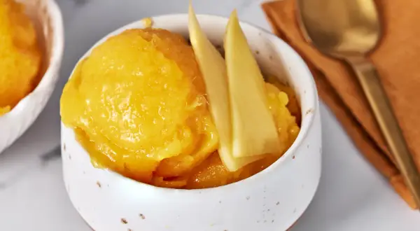 Erfrischendes Mango Sorbet in einer Schale, garniert mit Mangostücken. Daneben ein Löffel auf einem orangen Tuch.