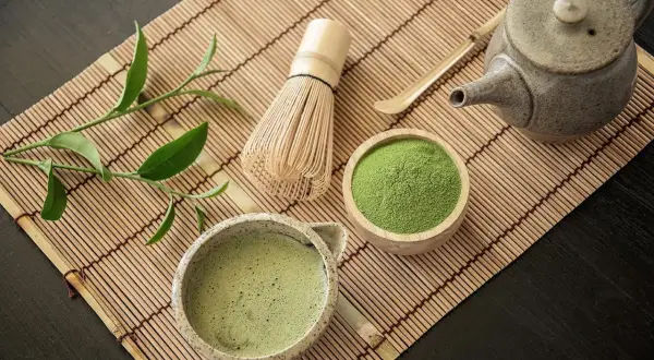 Tradycyjne akcesoria do parzenia matcha: bambusowa miotełka, czarka z napojem, drewniana miseczka z zielonym proszkiem.