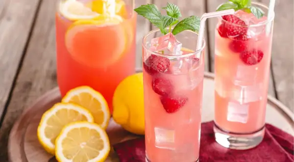 Två glas med rosa lemonad, hallon, is och mynta. Citroner och en karaff i bakgrunden ger härliga sommar vibes.