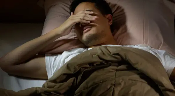 Man in bed, hand over eyes, can't sleep after long run. The runner's mind races, body tired but brain won't switch off.