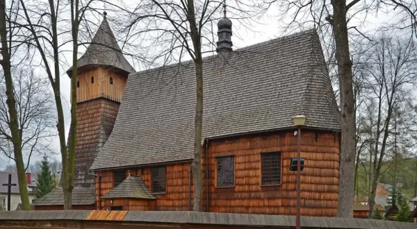 Drewniane kościoły południowej Małopolski: zabytkowa świątynia z wieżą i gontowym dachem, otoczona drzewami i drewnianym płotem.