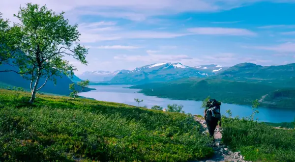 Vandra i Norrland med storslagen utsikt över sjö och snöklädda berg. En ensam vandrare med ryggsäck går på en stenstig.
