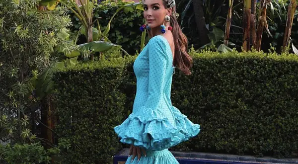 Una mujer luce un vestido de flamenca turquesa con lunares, adornado con volantes y una flor en la cabeza.