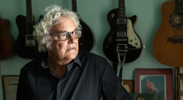 Massimo Bubola, con i suoi capelli ricci e bianchi, guarda pensieroso tra le chitarre appese al muro, testimoni silenziose delle sue canzoni.