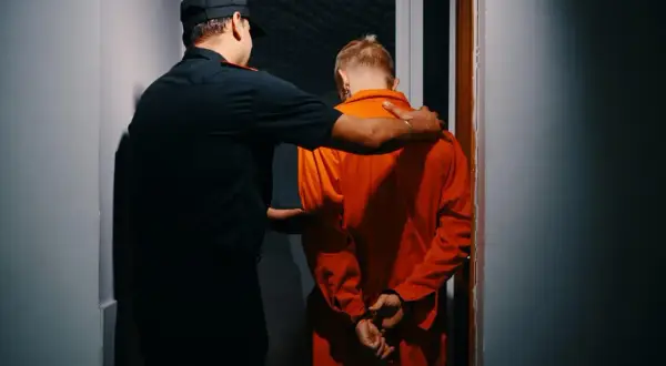 Un détenu en orange, menotté, est conduit par un gardien. La scène évoque une **garde à vue 48h**.