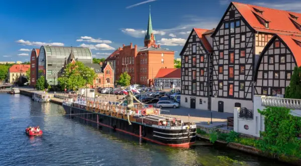 Nad rzeką w Bydgoszczy, jednym z 20 największych miast w Polsce, cumuje barka. W tle zabytkowe kamienice i nowoczesna architektura.