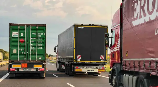 Trzy ciężarówki jadące autostradą. Znak zakazu wyprzedzania dla ciężarówek jest widoczny, sugerując ograniczenia ruchu.