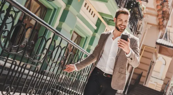 Ein Mann im casual chic Outfit mit Sakko und Jeans lächelt, während er auf sein Smartphone schaut.
