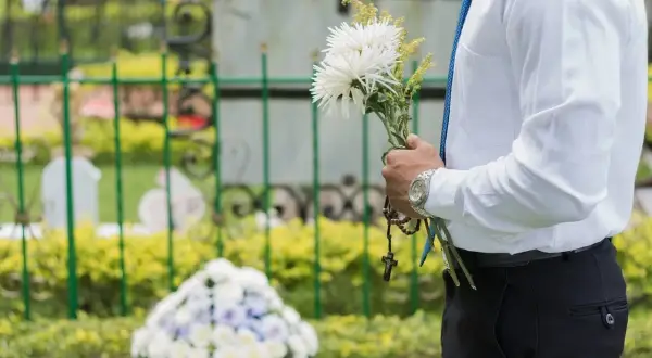 Mężczyzna w białej koszuli i niebieskim krawacie trzyma bukiet białych kwiatów i różaniec. W tle widać nagrobki i zielone krzewy. Msza pogrzebowa z urną trwa.