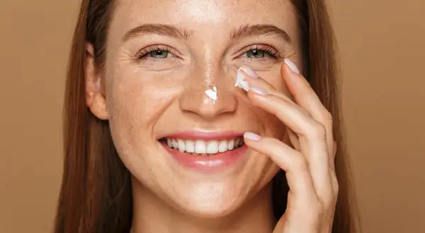 Uśmiechnięta kobieta z piegami aplikuje krem na nos. Nawet latem można stosować retinol, dbając o skórę.