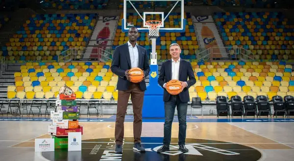 Dos hombres posan con balones de baloncesto en una cancha vacía. Detrás de ellos, una pila de cajas de frutas y asientos de colores. Un guiño al club baloncesto Gran Canaria.