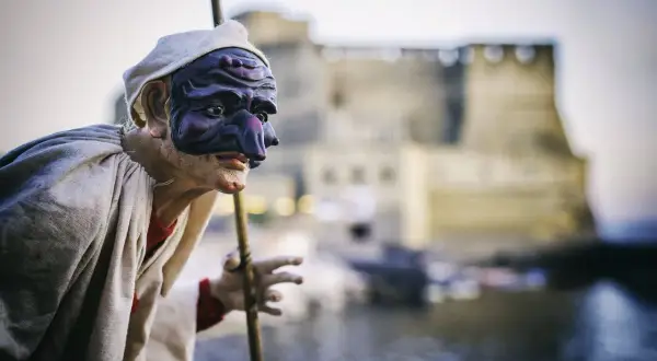 Pulcinella con maschera viola, pronto a litigare in napoletano, con Castel dell'Ovo sullo sfondo.