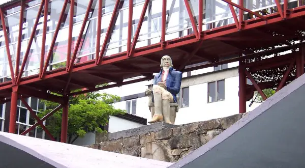 Estatua de un hombre sentado frente a la fábrica de Sargadelos, bajo un puente rojo y acristalado.